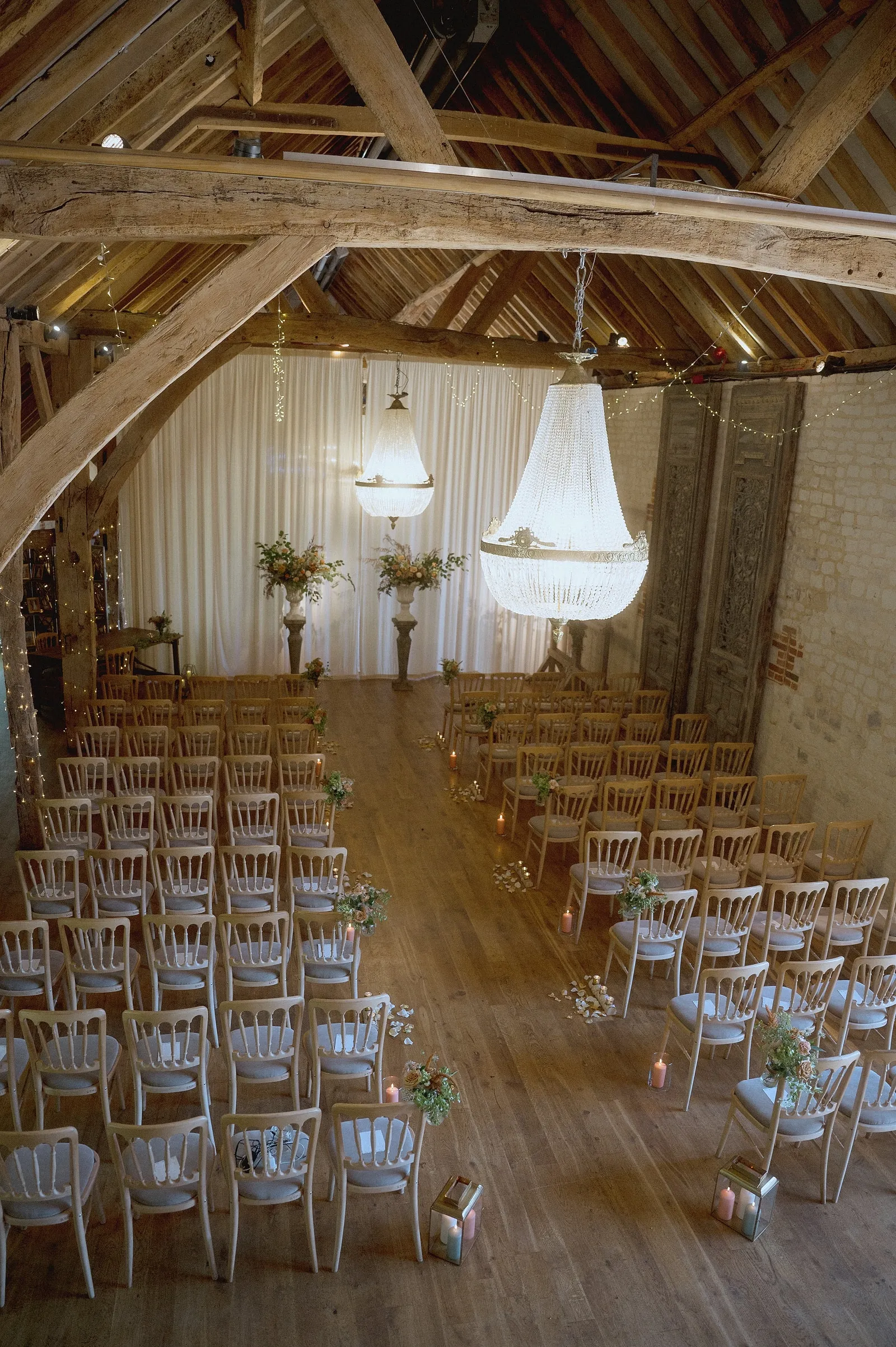The barn set for a ceremony: chairs either side of the aisle, crystal chandeliers under the oak beams, candles and flowers
