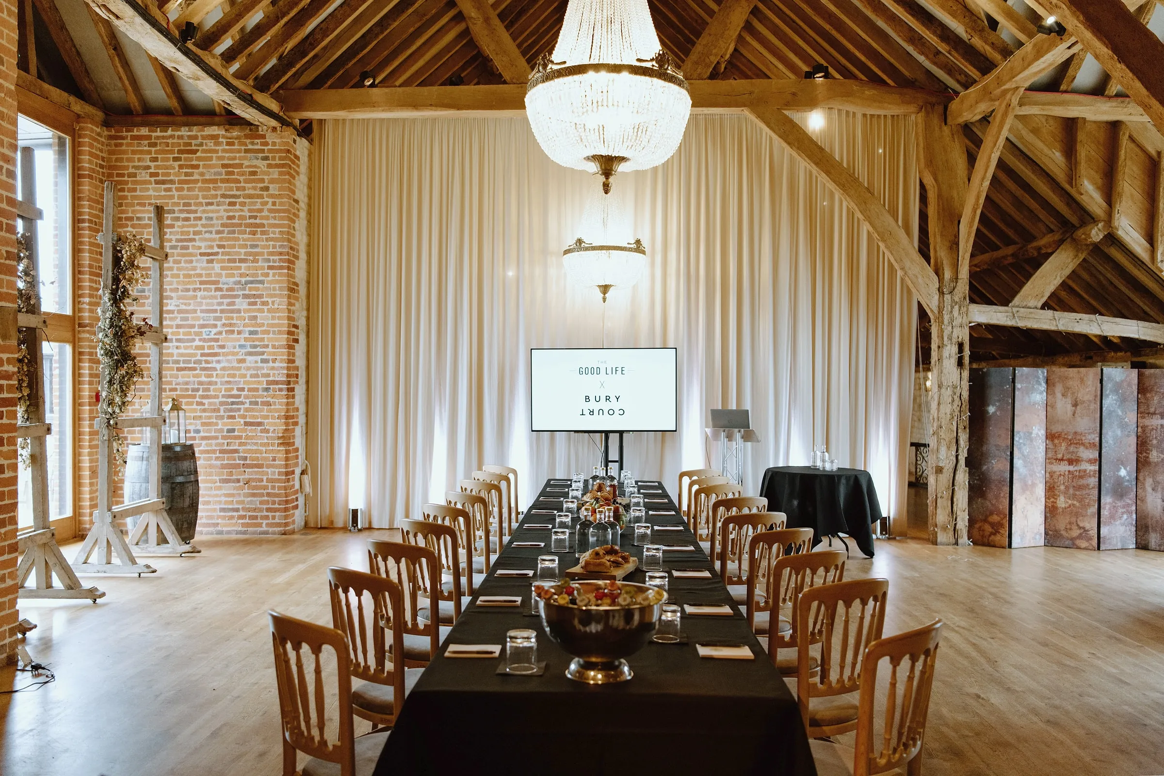 A long table set for a meeting under crystal chandeliers in the barn, with a screen and lectern at the far end
