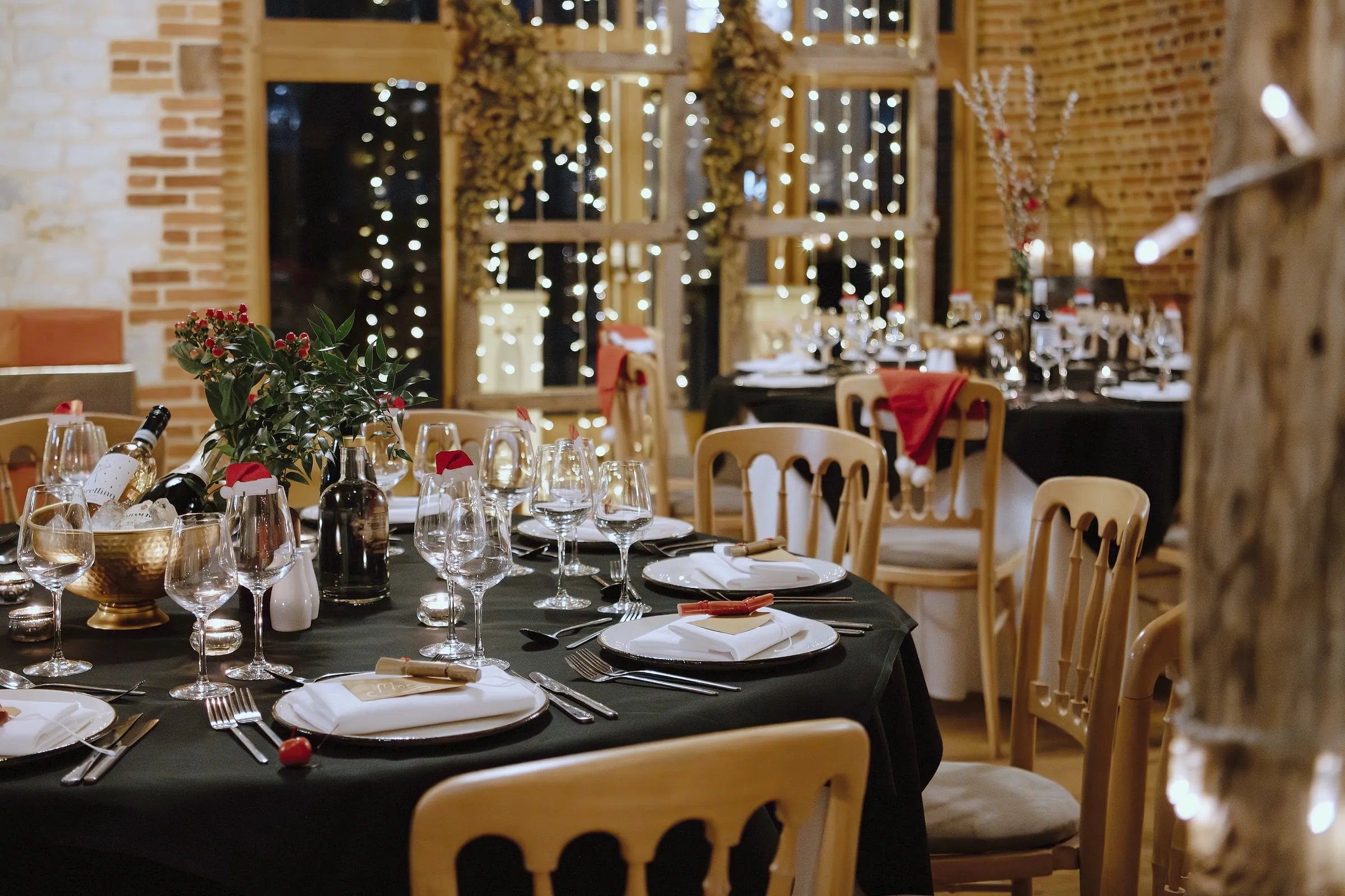Round tables in the barn laid for a festive dinner with black linen, glassware and fairy lights