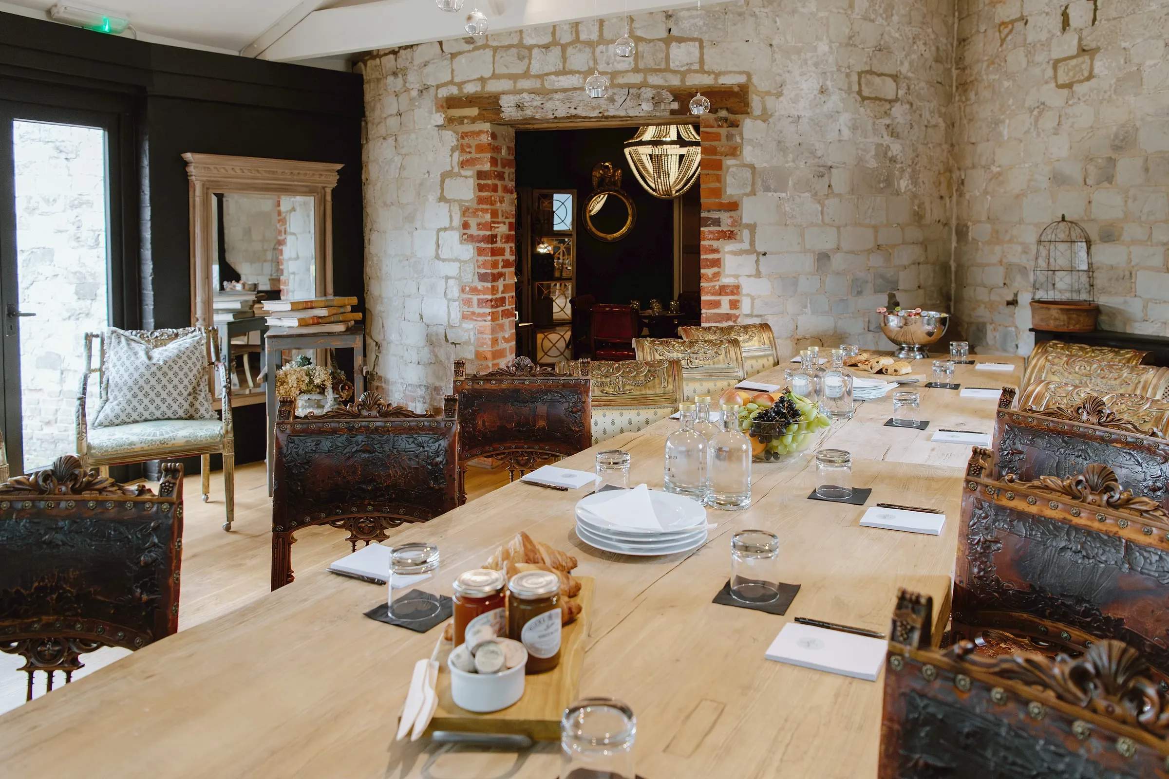 A long oak table laid for a meeting with carved chairs, notepads and pastries, in a stone room with a chandelier