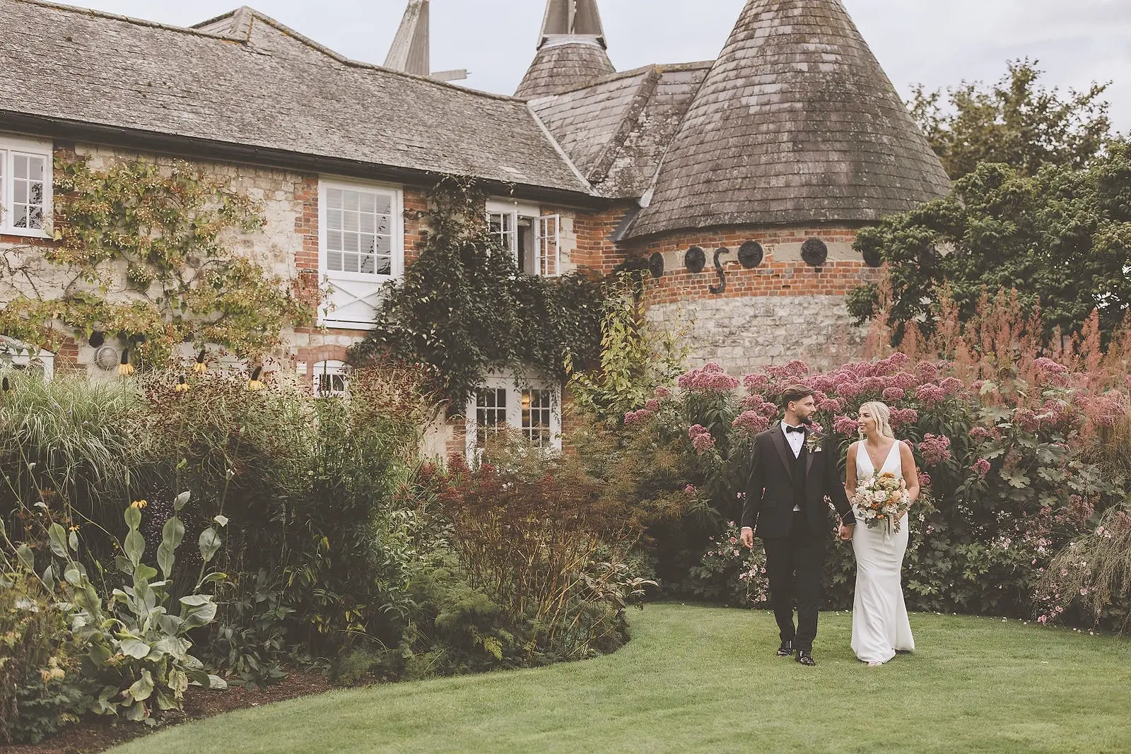 A couple walk hand in hand across the lawn past the oast houses, the borders in full late-summer flower