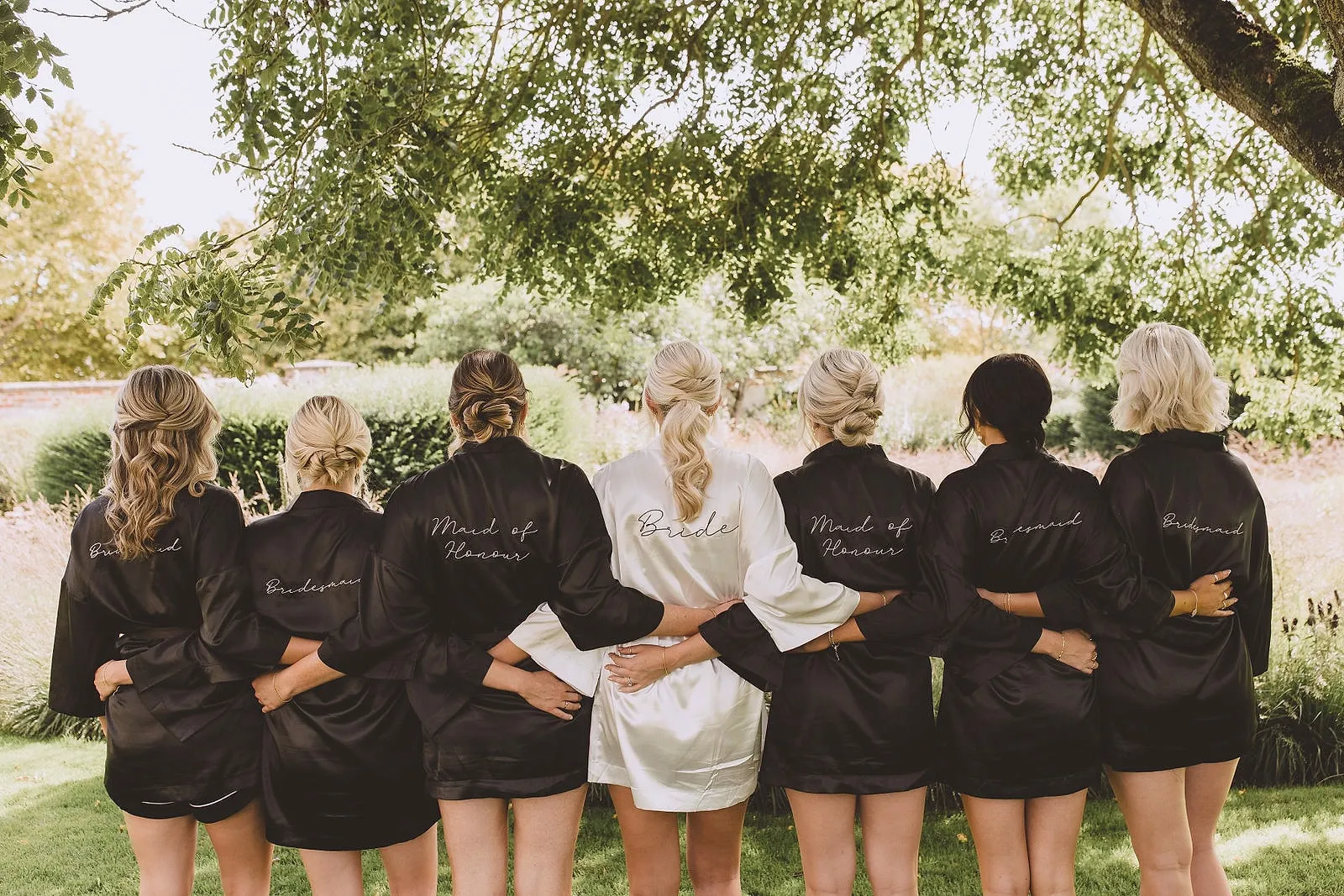 A bride in a white robe and her bridesmaids in black, arms round each other, facing the garden under a tree